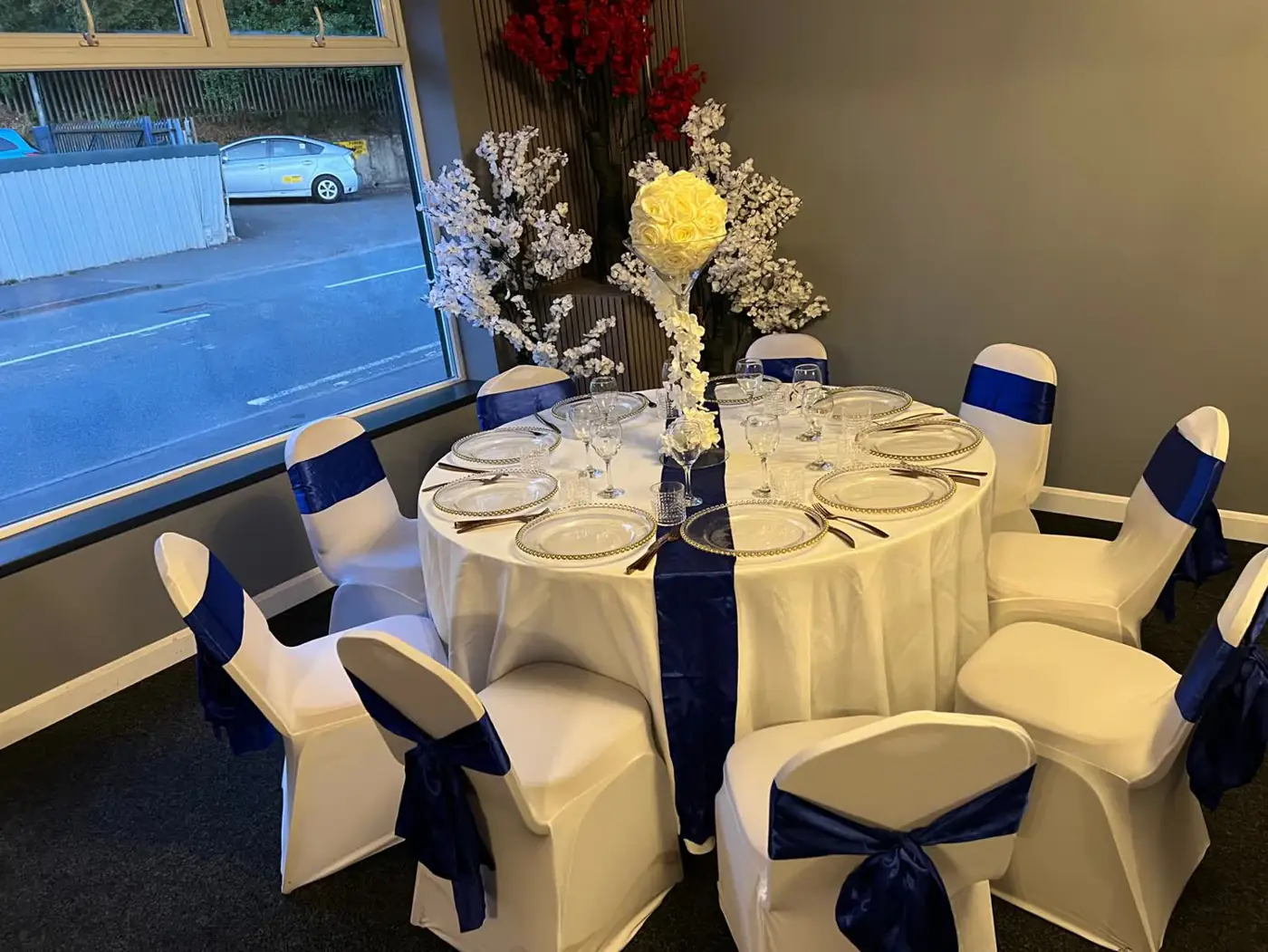 Silver Hall round table with a navy runner, navy chair sashes and a tall white floral centrepiece beside the window
