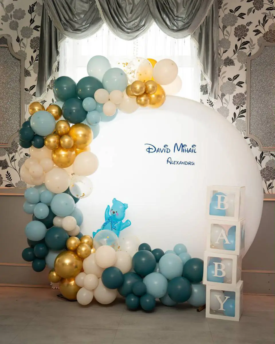 Sage green, teal and gold balloon arch around a white backdrop with 'BABY' letter blocks