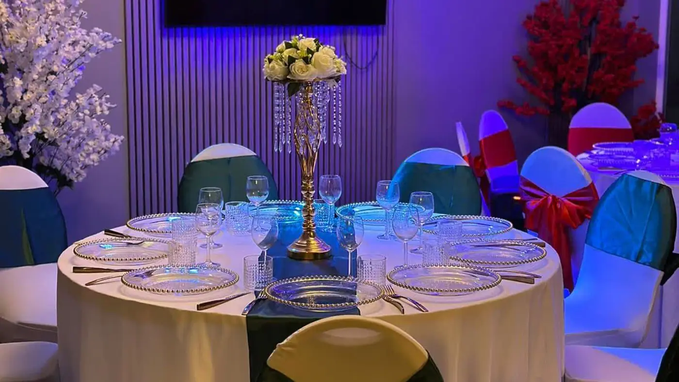 Table set under purple lighting with a tall crystal centrepiece, white blossom and teal and red chair covers